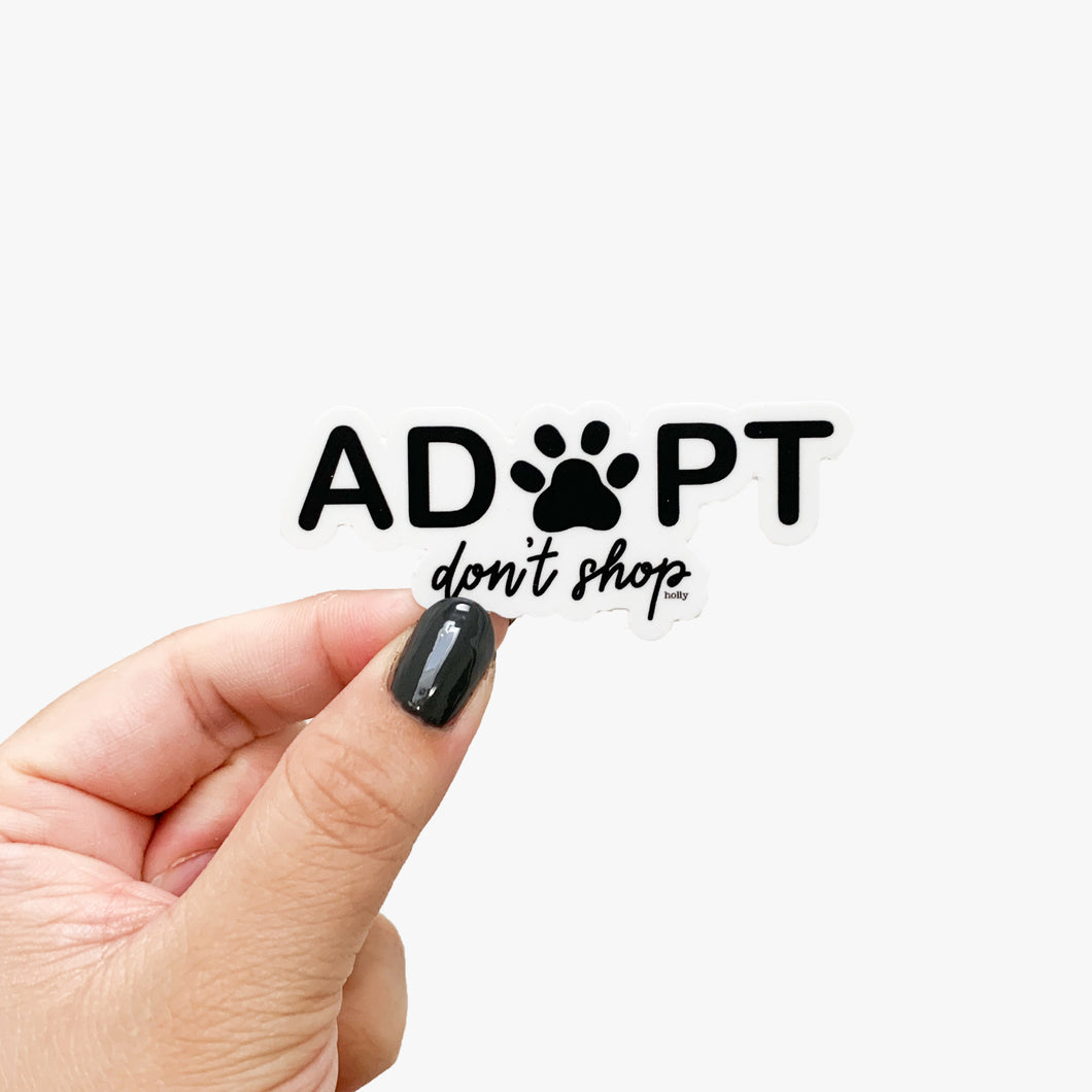 A black and white sticker with text that reads, Adopt don’t shop, and a paw print as the letter “O” in “Adopt”.