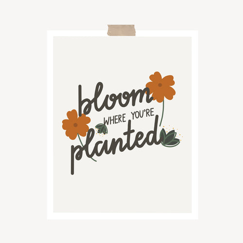 An art print featuring the popular quote, “Bloom where you’re planted,” written in calligraphy and decorated with orange flowers and green leaves hung with a piece of kraft tape on the top.