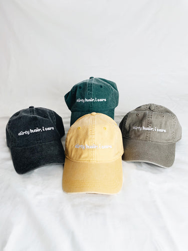 Four vintage washed baseball caps in black, green, yellow, and khaki with text on them that reads, Dirty hair, I care.