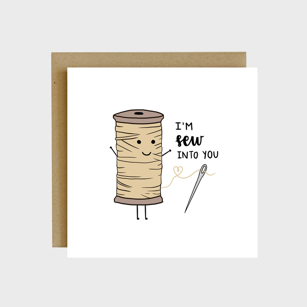 A funny greeting card featuring a sewing thread spool with a loose string in the shape of a heart that is threaded through a needle and text that reads, I’m sew into you.